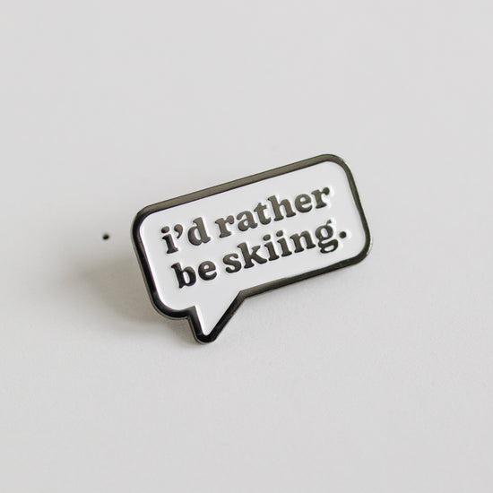 Enamel pin with 'i'd rather be skiing.' text on a light gray background