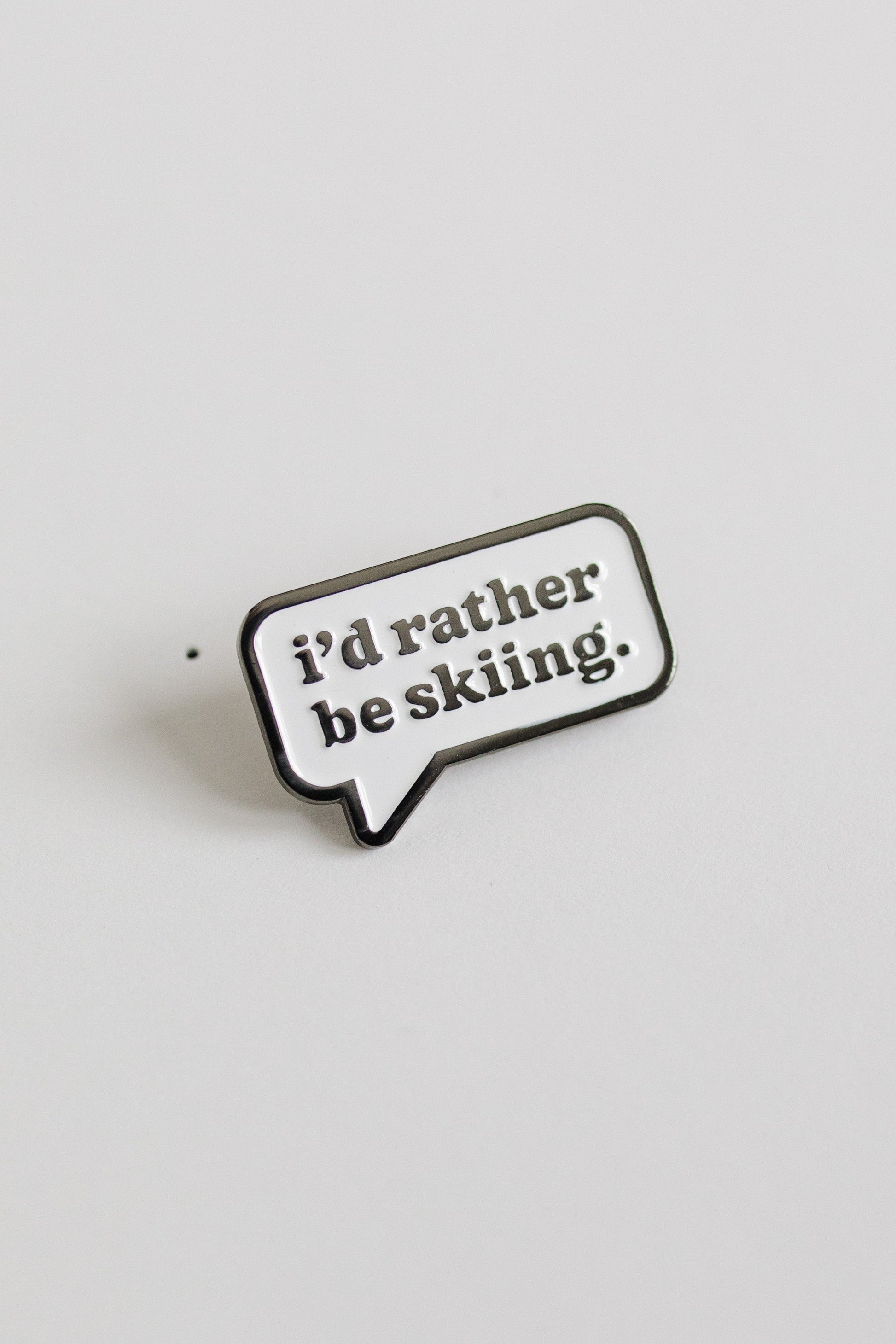 Enamel pin with 'i'd rather be skiing.' text on a light gray background