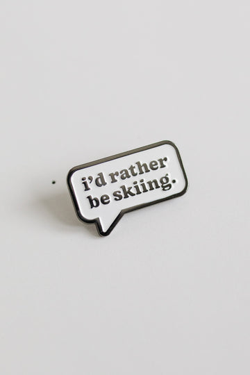 Enamel pin with 'i'd rather be skiing.' text on a light gray background