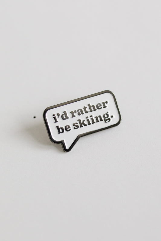 Enamel pin with 'i'd rather be skiing.' text on a light gray background
